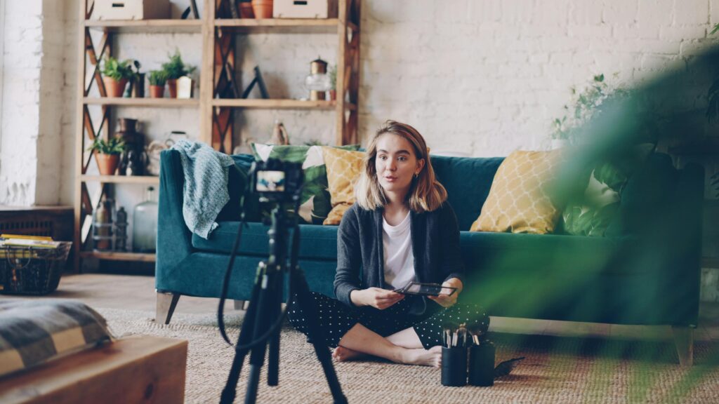 Canal no YouTube Sem Aparecer: Como Criar, Monetizar e Ganhar Dinheiro com o Método Dark Woman filming vlog at home with camera setup, surrounded by plants and cozy decor.