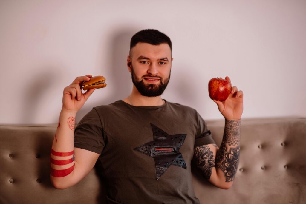 Disciplina x Motivação: Por Que Uma Funciona e a Outra Falha Bearded man pondering diet choice with apple and burger. Indoors with tattoos visible.