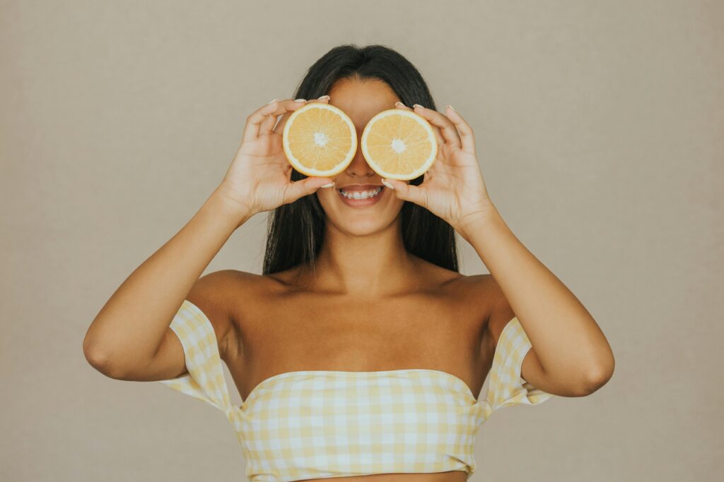 Pele Saudável Começa Por Dentro: O Que Comer Para Ter Boa Pele Smiling woman with long hair playfully holds orange halves over her eyes.