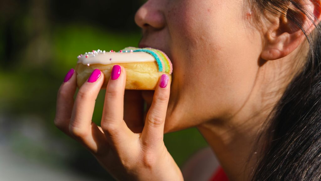 O Que Acontece Com Seu Corpo Quando Você Para de Comer Açúcar Por 30 Dias A woman with manicured nails takes a bite of a colorful sprinkled doughnut.