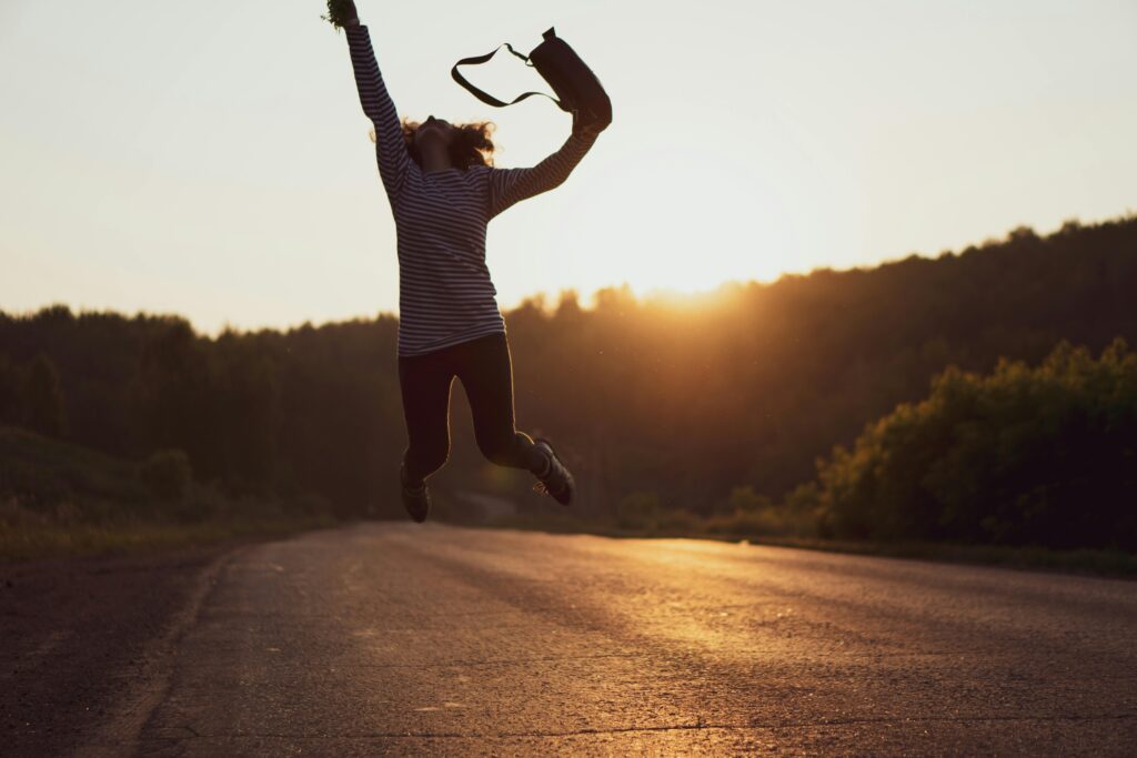 Como Vencer a Ansiedade com Técnicas Clínicas Comprovadas Silhouette of a woman jumping joyfully on an open road at sunset, embodying freedom and adventure.