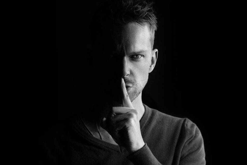 Como Se Afastar de Pessoas Narcisistas Sem Drama e Sem Culpa Black and white portrait of a man gesturing for silence, highlighting mystery.
