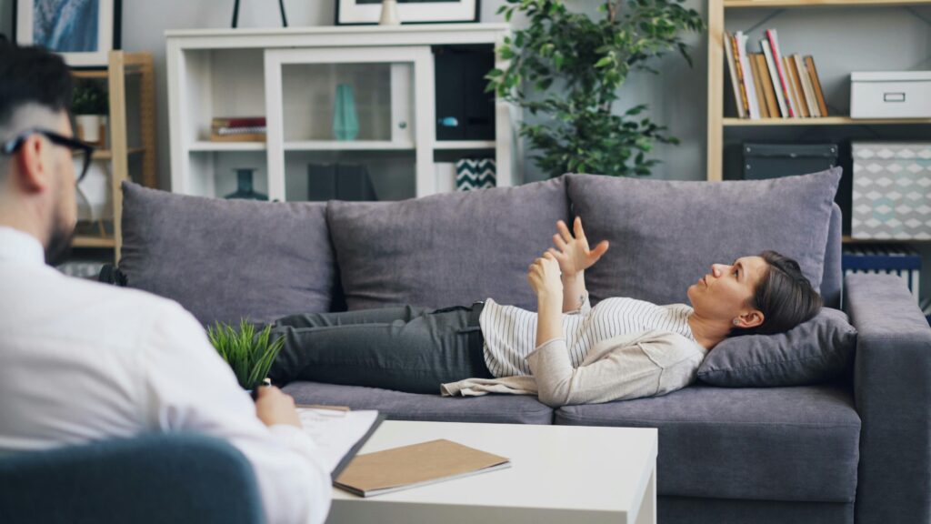 Como Cuidar da Saúde Mental No Trabalho Remoto A woman in therapy, speaking with a psychologist while lying on a sofa, indoors.