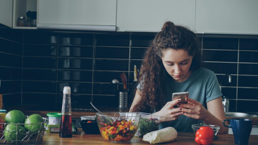 Alimentos Que Melhoram a Concentração e o Foco (Baseado em Ciência) A young woman checks her smartphone in a modern kitchen with fresh vegetables.