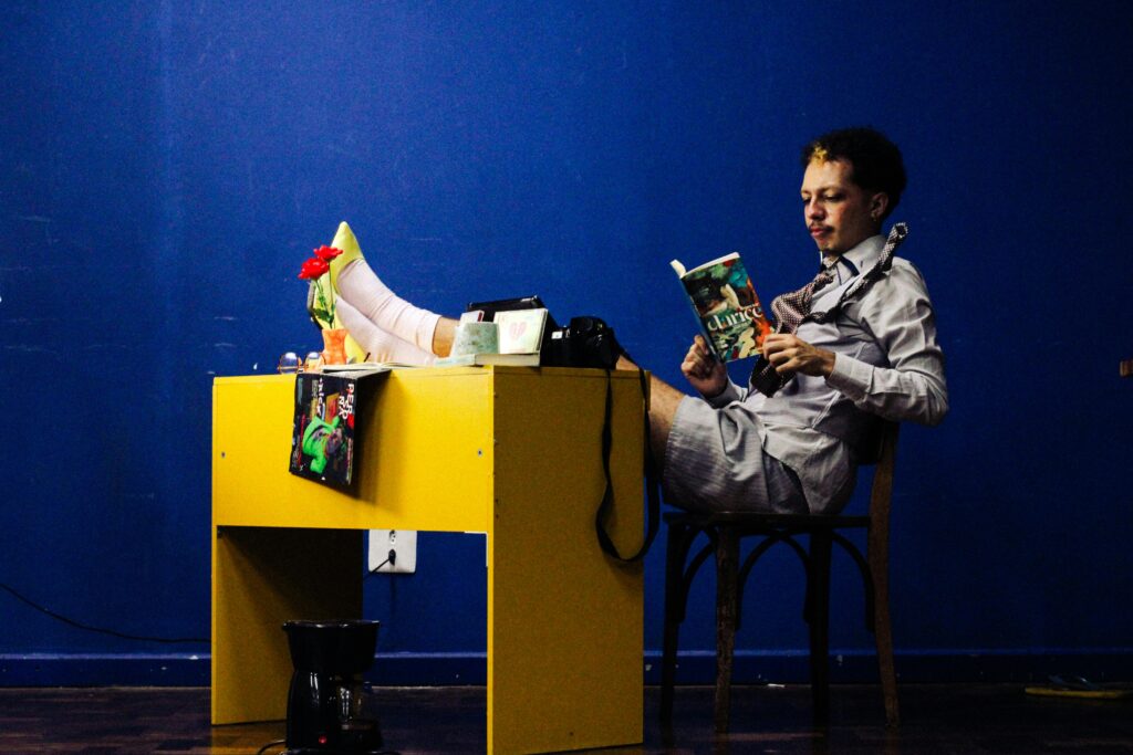 Man with a leg cast reading a book at a yellow desk in a blue-walled room, exuding a relaxed indoor atmosphere.
