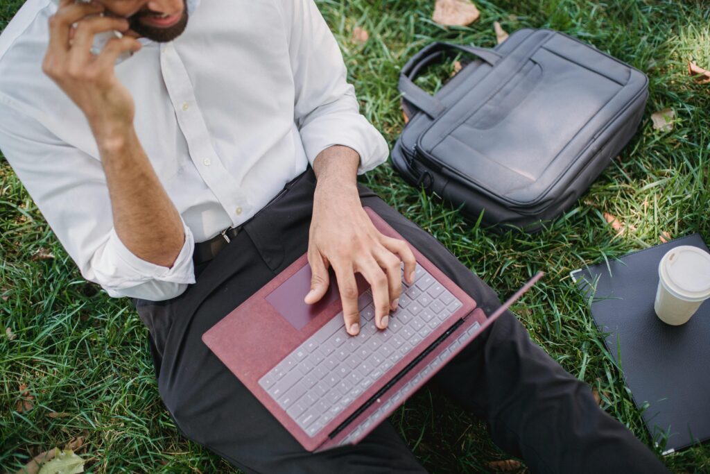 A businessman in formal attire working on a laptop while sitting on grass in a park setting.