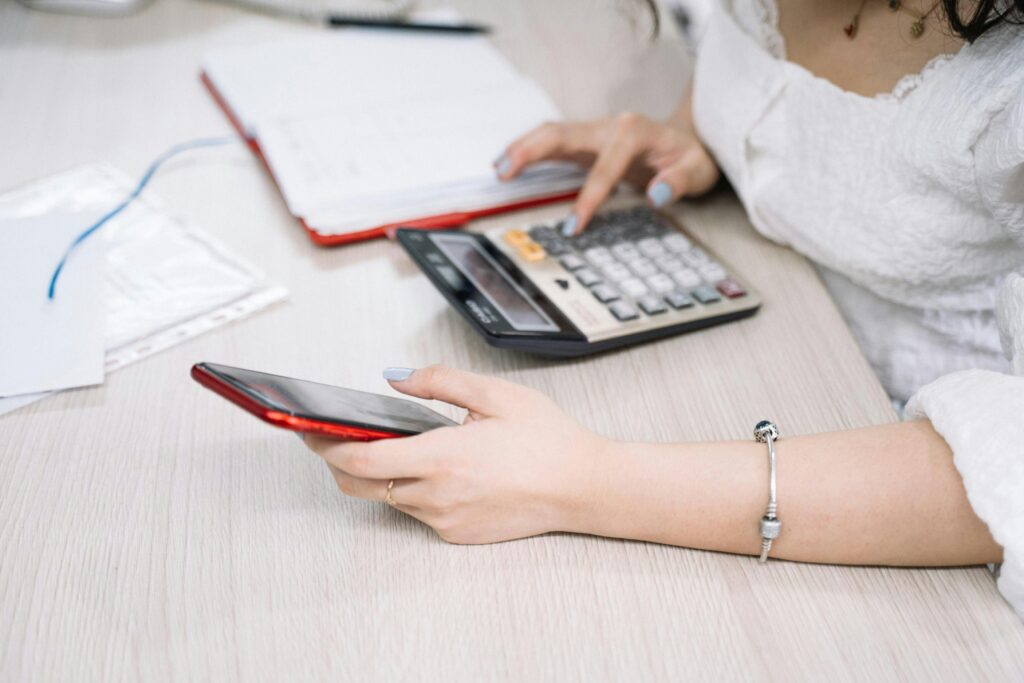 Como Precificar Seus Serviços Digitais Sem Medo de Perder Clientes A woman managing finances using a calculator and smartphone at her desk, showcasing multitasking in an office setting.