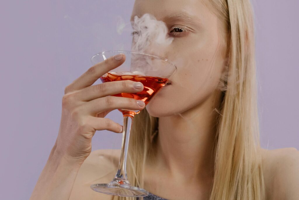 O Impacto do Álcool no Corpo: O Que Acontece Depois de Apenas Uma Taça A woman savoring a vibrant, smoky cocktail in a martini glass with blonde hair indoors.