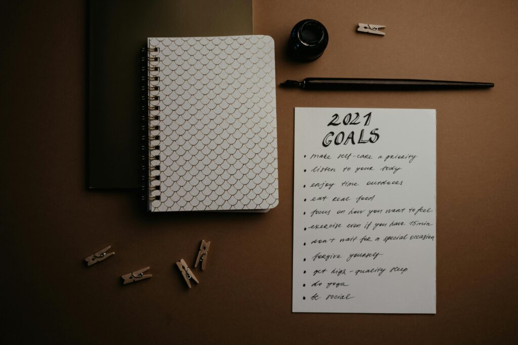 Como Criar Metas Que Você Realmente Vai Cumprir Aesthetic flatlay of notebooks and a 2021 goals list in elegant calligraphy.