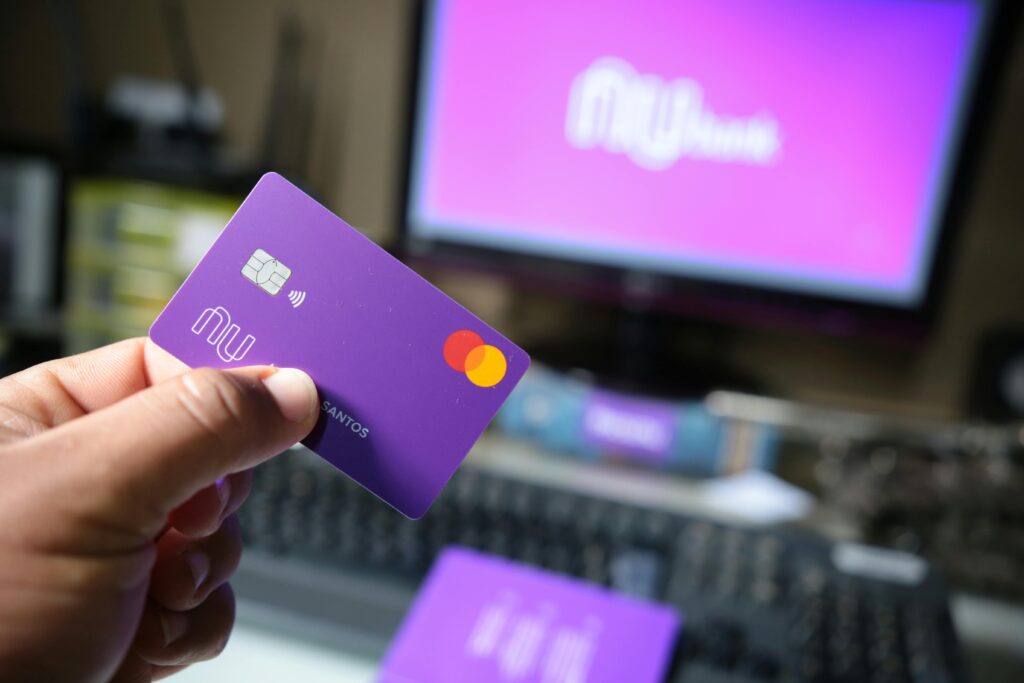 Como Usar o Nubank, Inter ou C6 Para Organizar Suas Finanças A close-up of a hand holding a purple bank card in front of a computer screen.