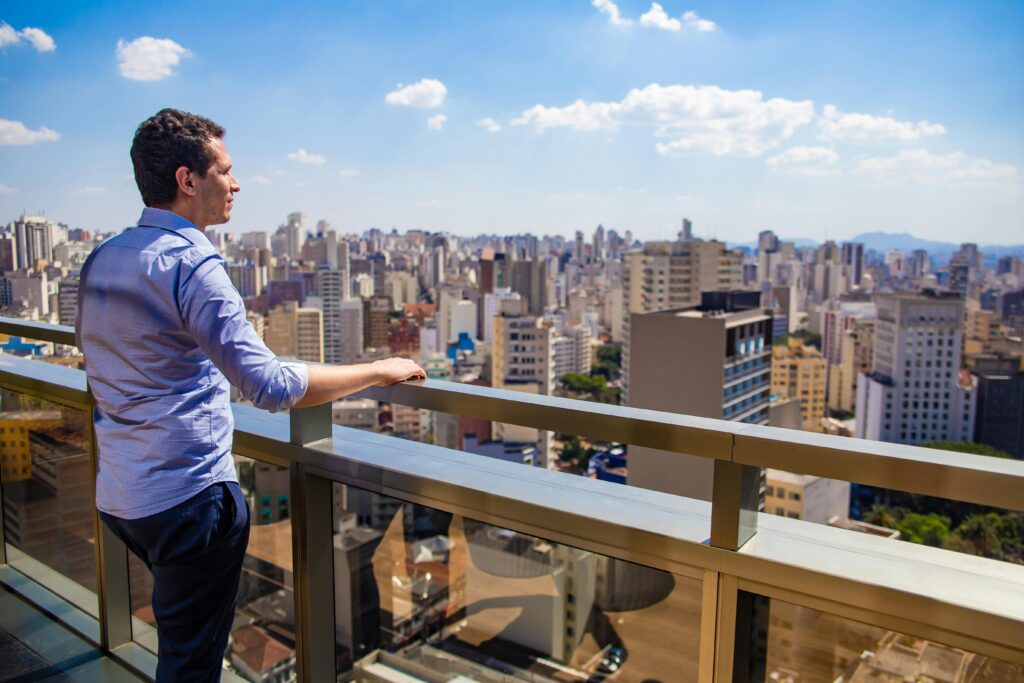 Como Trabalhar Para Empresas Estrangeiras Morando no Brasil A man in a shirt stands on a terrace, looking at the São Paulo cityscape under a clear day sky.