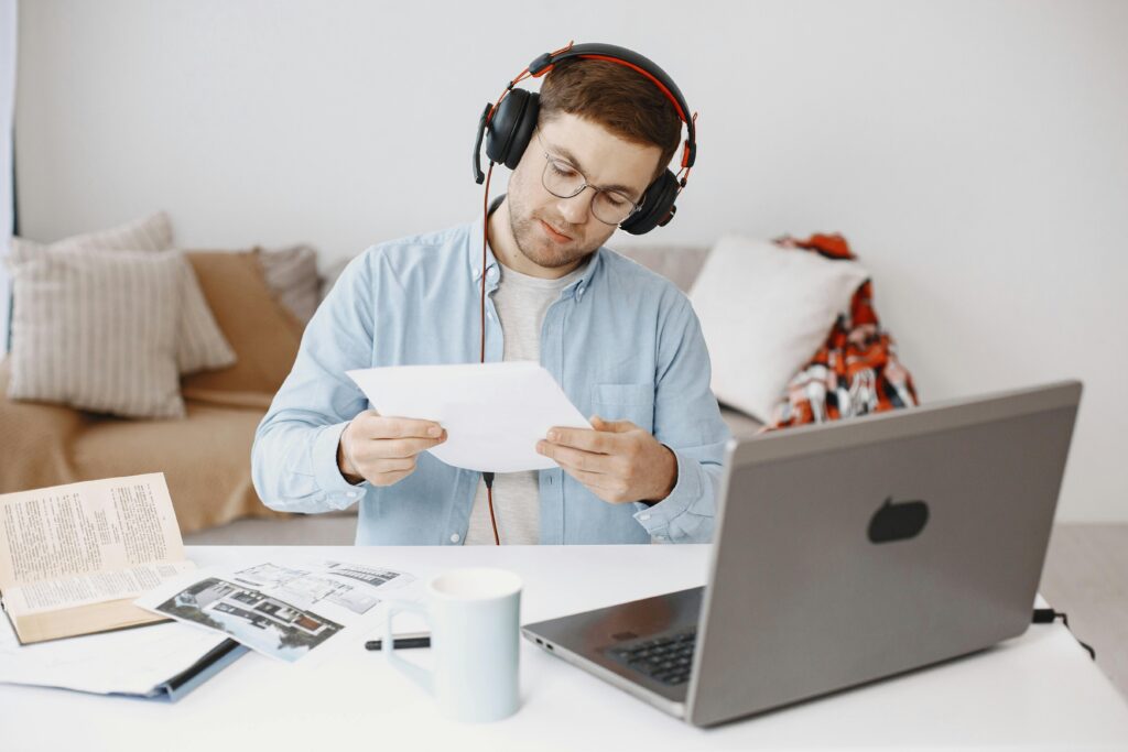 Adult man focused on documents while working remotely at home with laptop and headphones.