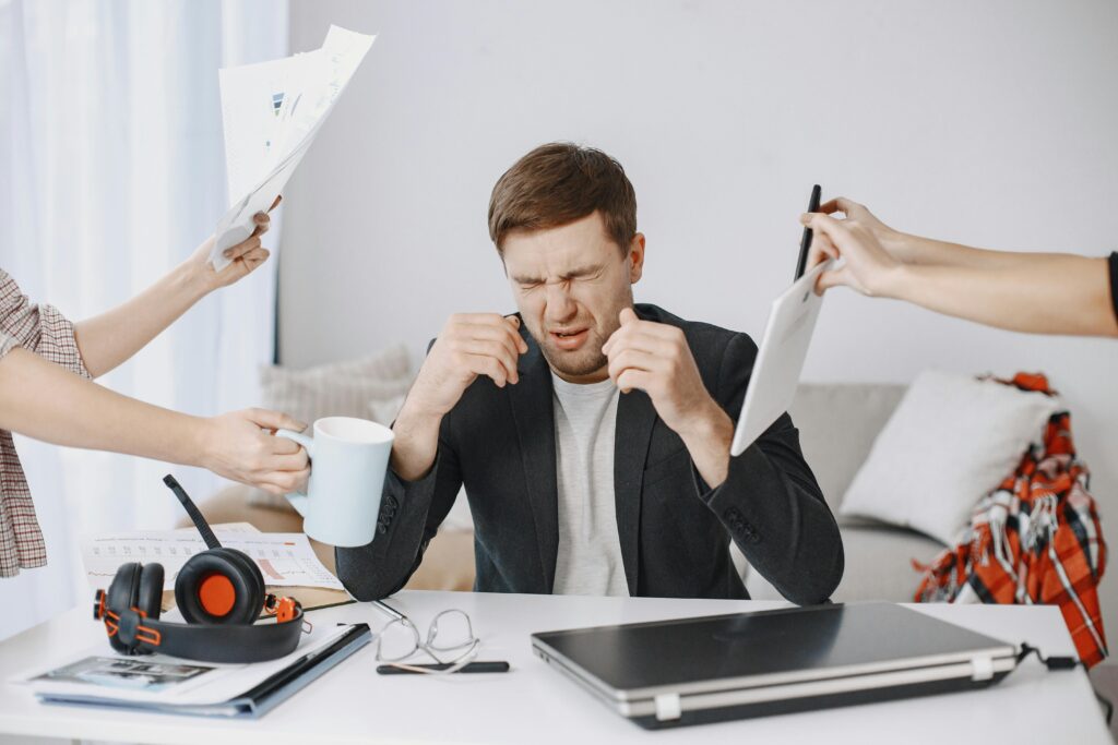Como Criar Metas Que Você Realmente Vai Cumprir Adult man stressing out at home office desk with multiple demands surrounding him.