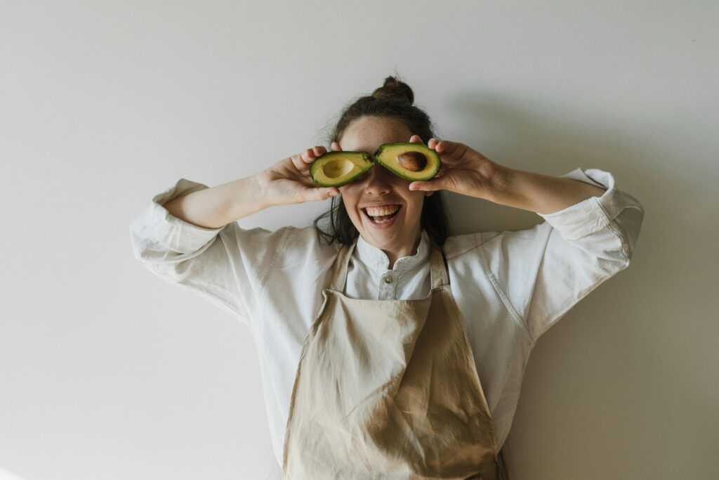 Como a Alimentação Influencia Diretamente Seu Humor e Sua Energia Joyful woman holding avocado halves over her eyes, symbolizing healthy living and fun creativity.