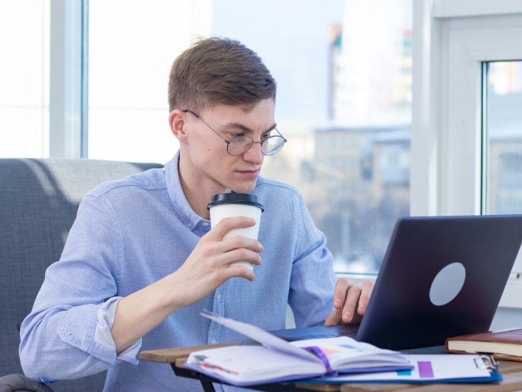Como Ser um Freelancer de Sucesso Sem Ter Experiência Anterior Adult wearing eyeglasses works remotely on a laptop while enjoying coffee indoors.