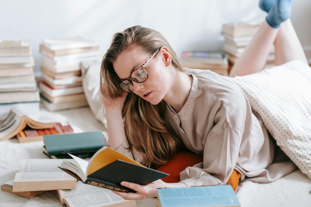 O Hábito de Ler Pode Mudar Sua Vida? A Ciência Responde Concentrated female in eyeglasses reading book while lying on floor with plaid and collection of literature in light room at home