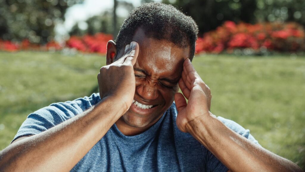 O Papel do Cortisol Na Sua Saúde: Por Que o Estresse Engorda Adult man outdoors with hands on head showing signs of discomfort or headache.