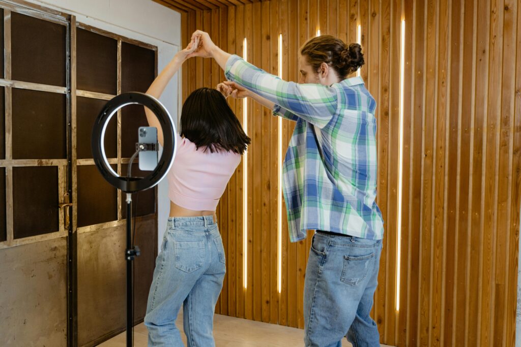 Como Ganhar Dinheiro com o TikTok Mesmo Sem Ser Famoso A young couple dances indoors with a ring light recording them, capturing a lively moment.