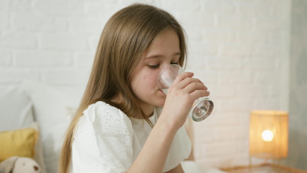 O Que Acontece com Seu Corpo Quando Você Bebe Água em Jejum Todo Dia A young girl drinks water in a cozy bedroom, highlighting hydration and comfort.