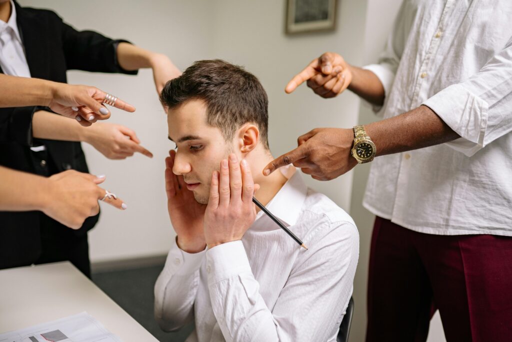 Como Identificar Uma Pessoa Narcisista (Antes Que Seja Tarde) A stressed man at the office surrounded by pointing fingers, representing workplace bullying and stress.