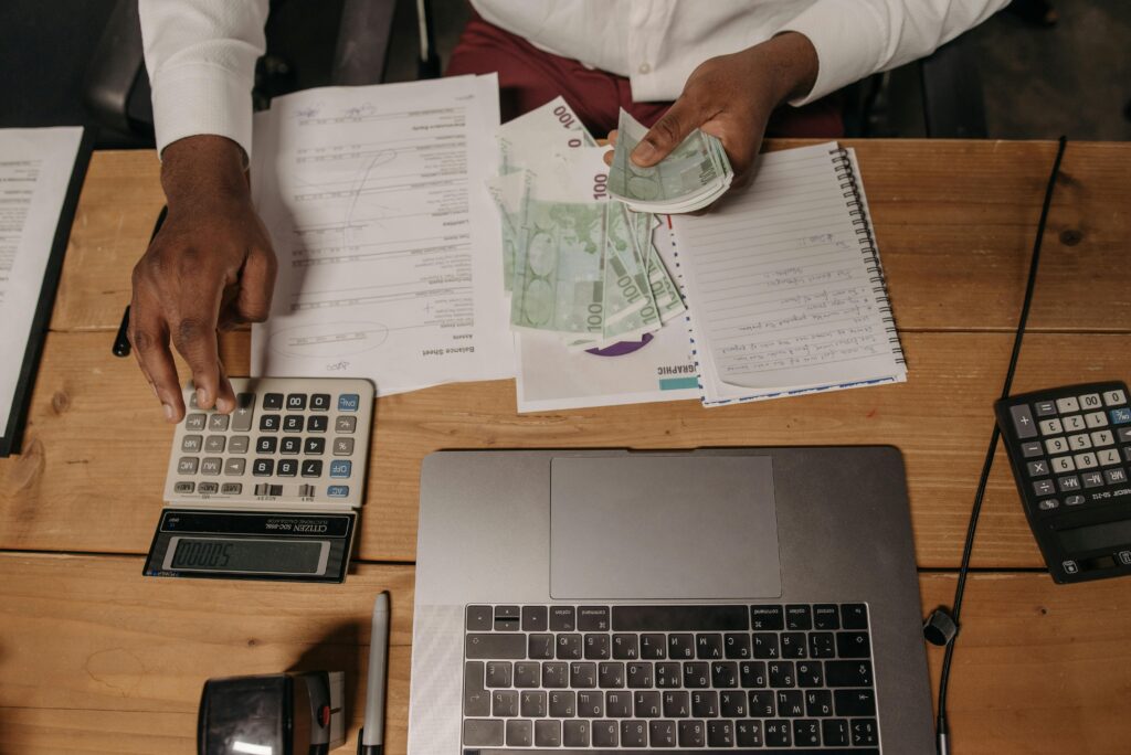 Como Definir Suas Tarifas Como Freelancer e Parar de Cobrar Barato Close-up of a person counting cash with documents and a laptop in an office setting.