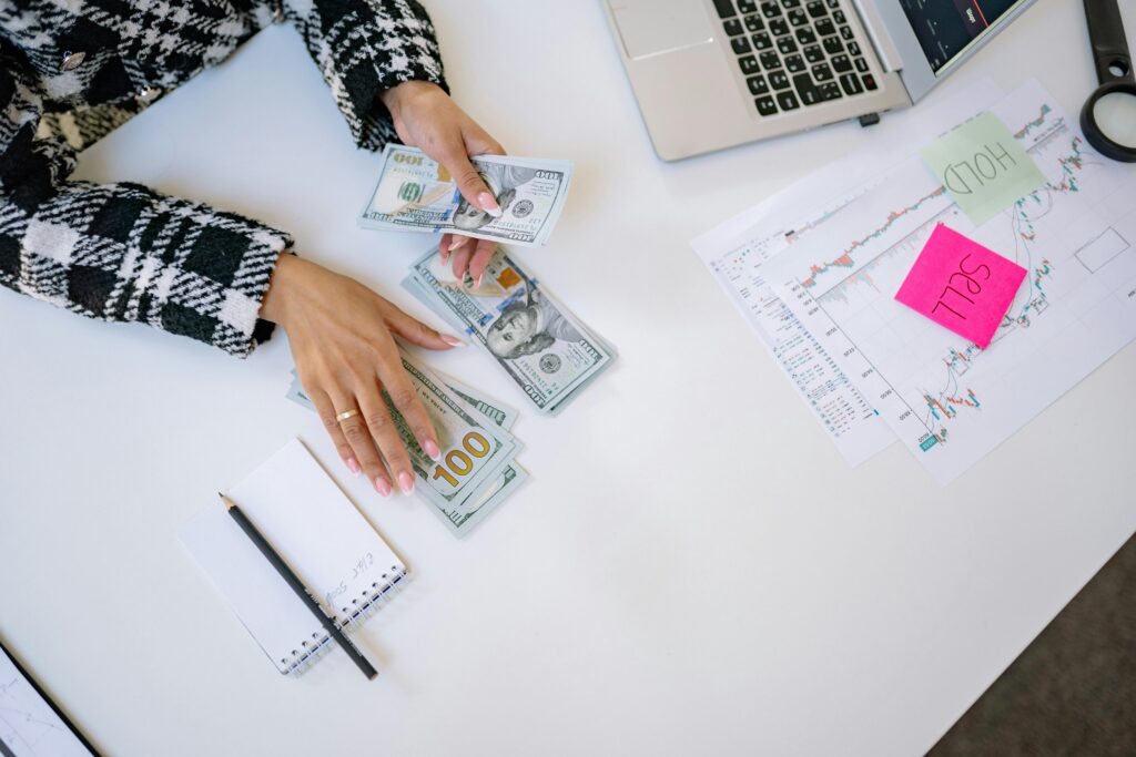 Como Investir Com Pouco Dinheiro: Começando com R$50 por Mês Businesswoman counting dollar bills with financial charts and laptop on table. Investment and finance concept.