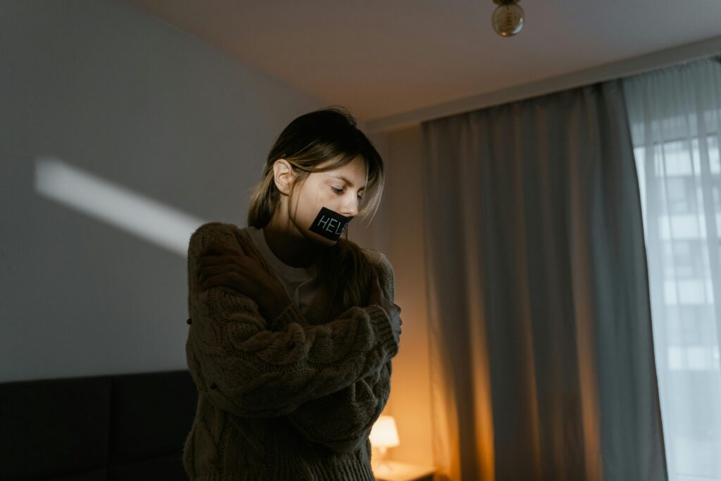 Gaslighting: O Que é e Como Saber Se Você Está Sendo Manipulado A young woman in a dim room wears a sweater and a help sign, expressing need.