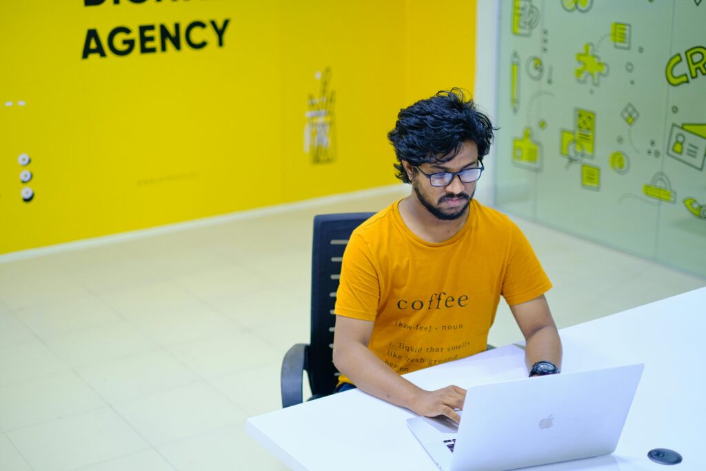 Como Montar Uma Agência Digital Solo e Faturar Alto Young man wearing eyeglasses working on laptop in vibrant digital agency office.