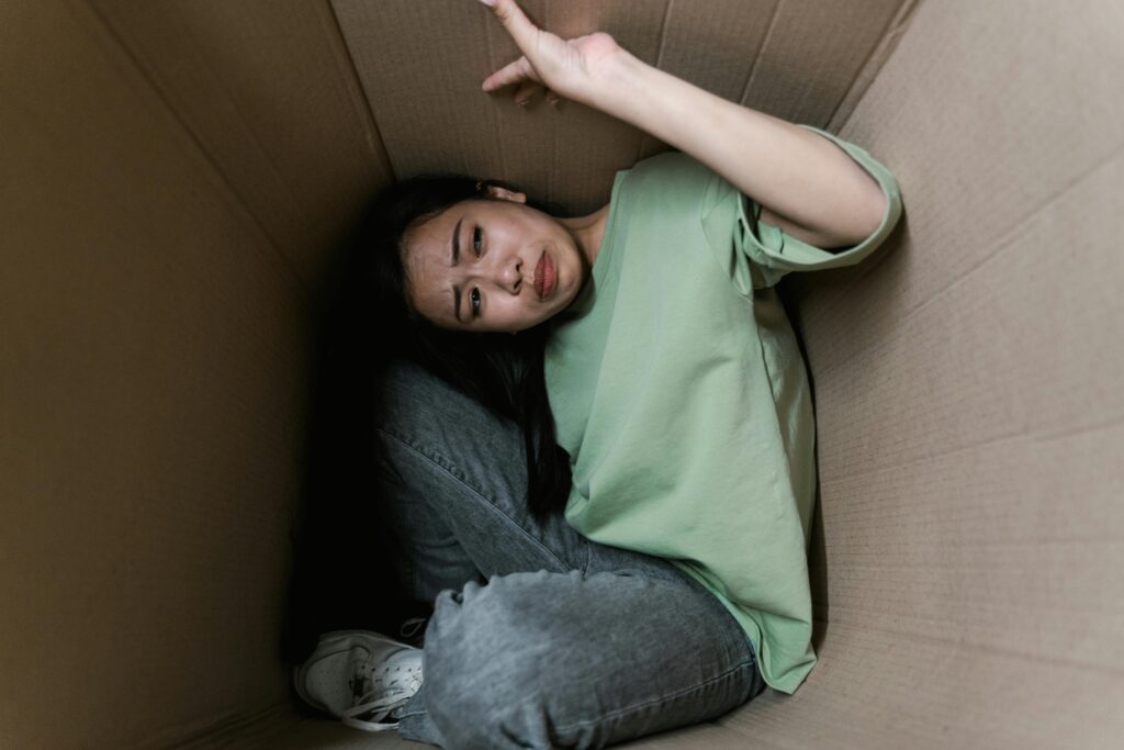 Como a Ansiedade Afeta Fisicamente Seu Corpo (Além da Cabeça) Woman enclosed in a cardboard box, showing signs of stress and fear.