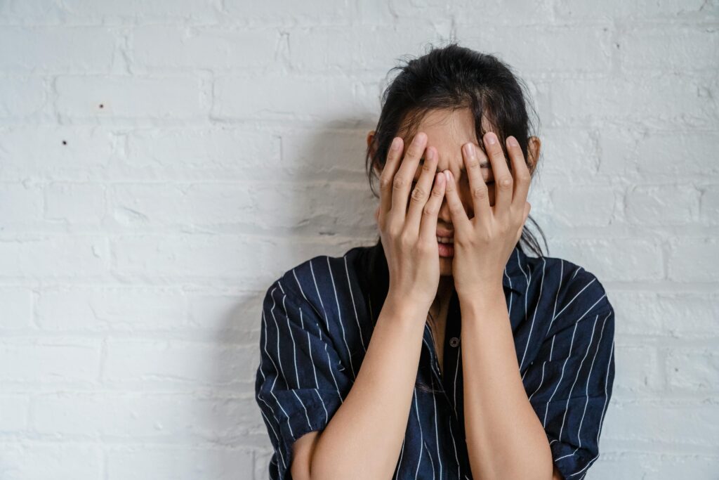 Superando a Ansiedade em 24 Dias: Um Método Natural e Comprovado A young woman with hands covering face expresses stress against a white brick wall background.