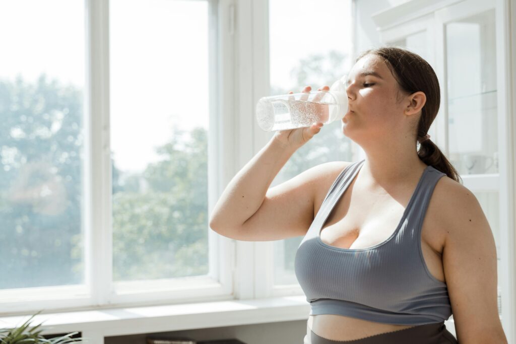 Hidratação: Quanto Água Você Realmente Precisa Beber Por Dia Active woman in sportswear drinks water indoors. Healthy lifestyle and hydration.