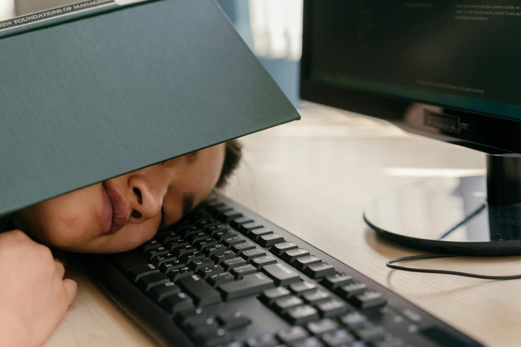 Como o Excesso de Tela Está Prejudicando Sua Visão e Seu Sono A young woman falls asleep on a keyboard with a textbook on her face, depicting study fatigue.