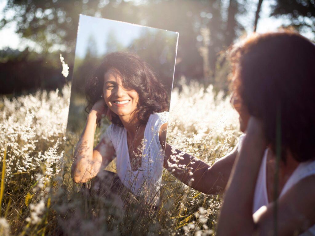 Desafio 21 Dias: Desbloqueie sua Melhor Versão com um Método Prático A woman smiles at her reflection in a mirror placed in a sunny field.