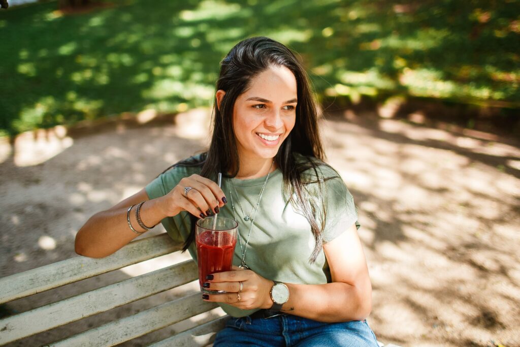 A smiling young woman enjoying a refreshing drink on a sunny day in the park.