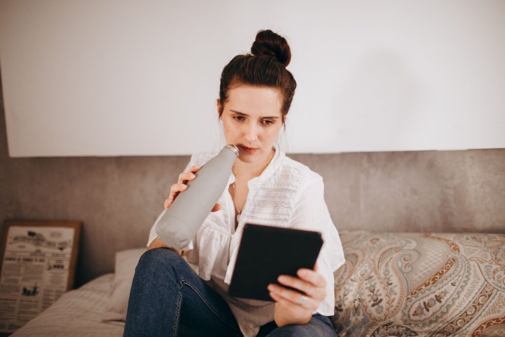 Young woman sitting on bed, reading an e-reader and drinking from a water bottle.