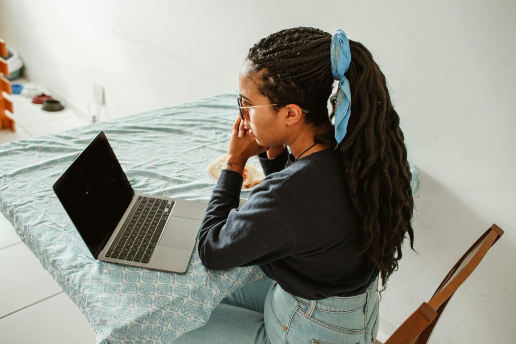 A young woman with braids and glasses studying on her laptop at a home table.