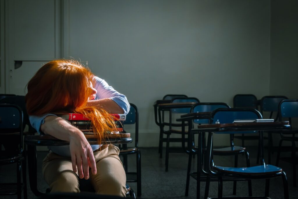 A tired student with red hair sleeps in an empty classroom under soft natural light.