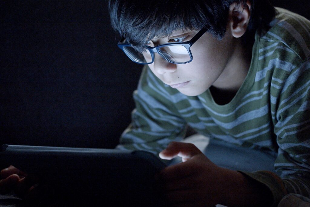 Young boy wearing glasses focused on tablet screen in dark room. Captures modern tech usage.