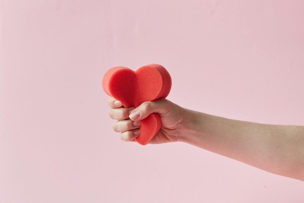Hand gripping a red foam heart against a pink background, symbolizing emotion and care.